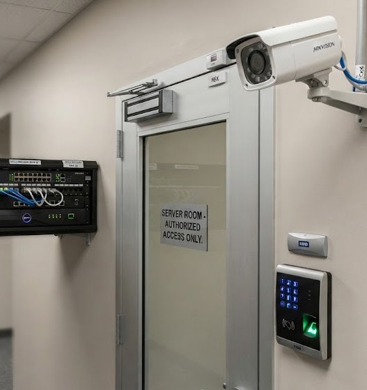 A server room door with an access control reader and keypad, a wall mounted comms cabinet and a CCTV camera covering the approach
